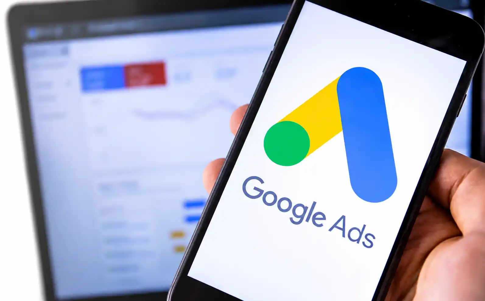 Phone screen shows Google Ads logo; a blurred analytics dashboard is in the background.